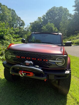 Red Ford Bronco with custom front winch and off-road accessories on display at Bronco Wild Fund Days, surrounded by trees and sunshine.