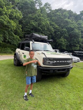 Smiling child posing in front of a white Ford Bronco at Bronco Wild Fund Days, surrounded by greenery and outdoor adventure vibes.