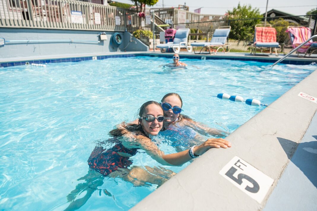 Two smiling girls swimming at a family-friendly hotel pool in Saugatuck Michigan, enjoying a sunny day at Beachway Resort with lounge chairs and summer vibes.