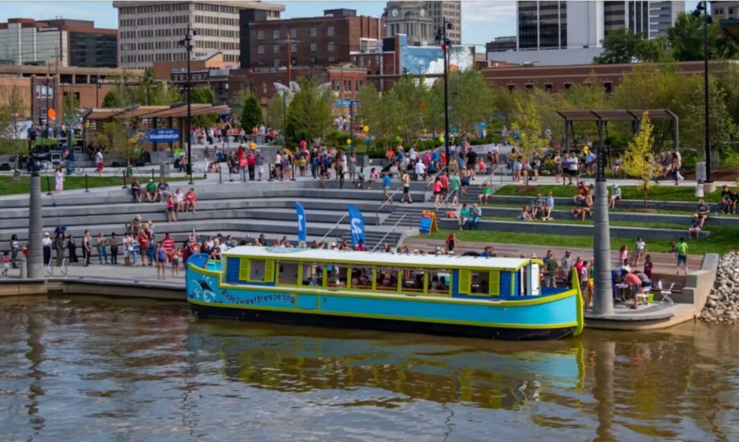 Crowds enjoy a sunny day at Promenade Park in Fort Wayne, Indiana, with the colorful Sweet Breeze canal boat docked by the riverfront.