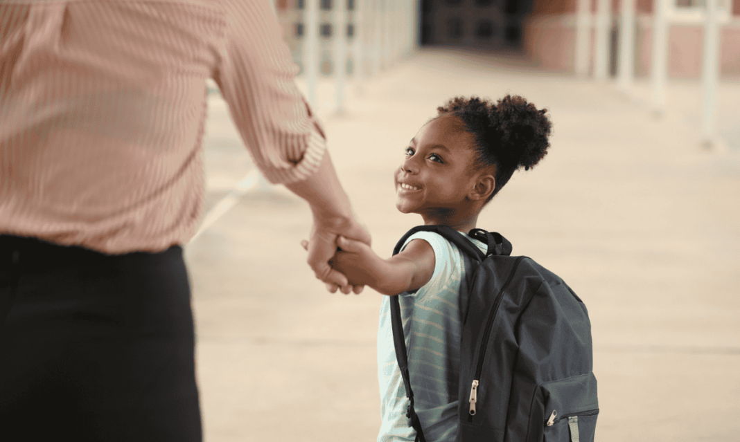 Smiling young student holding a parent’s hand while walking to school with a backpack.