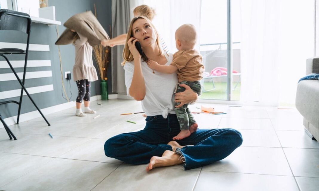 Parent sitting on the floor holding a baby while talking on the phone, with kids playing in the background, illustrating the need for lasting energy and healthy fuel for busy days.