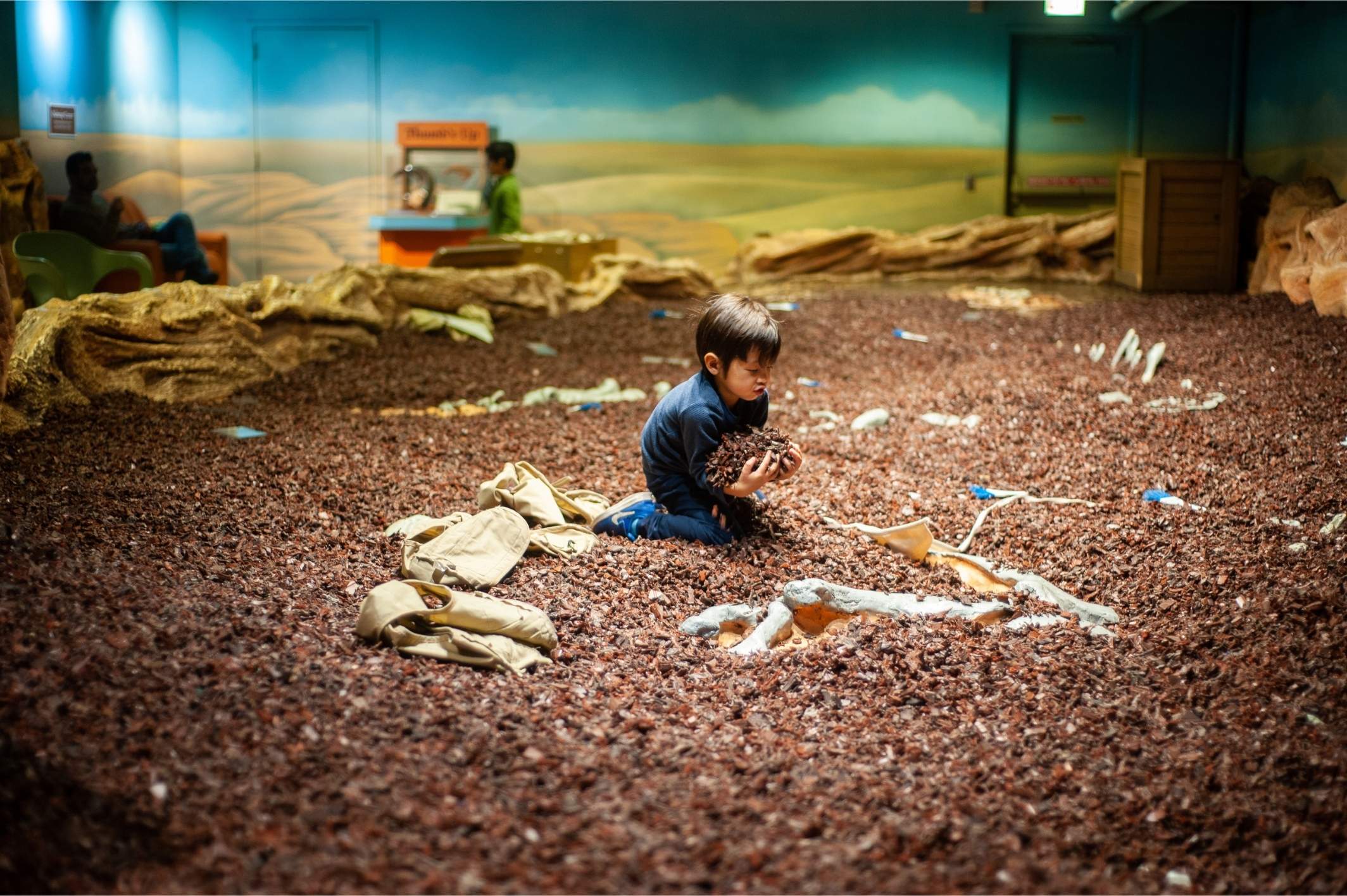 Young child plays in a fossil dig pit at the Chicago Children’s Museum, scooping bark chips while uncovering hidden dinosaur bones.