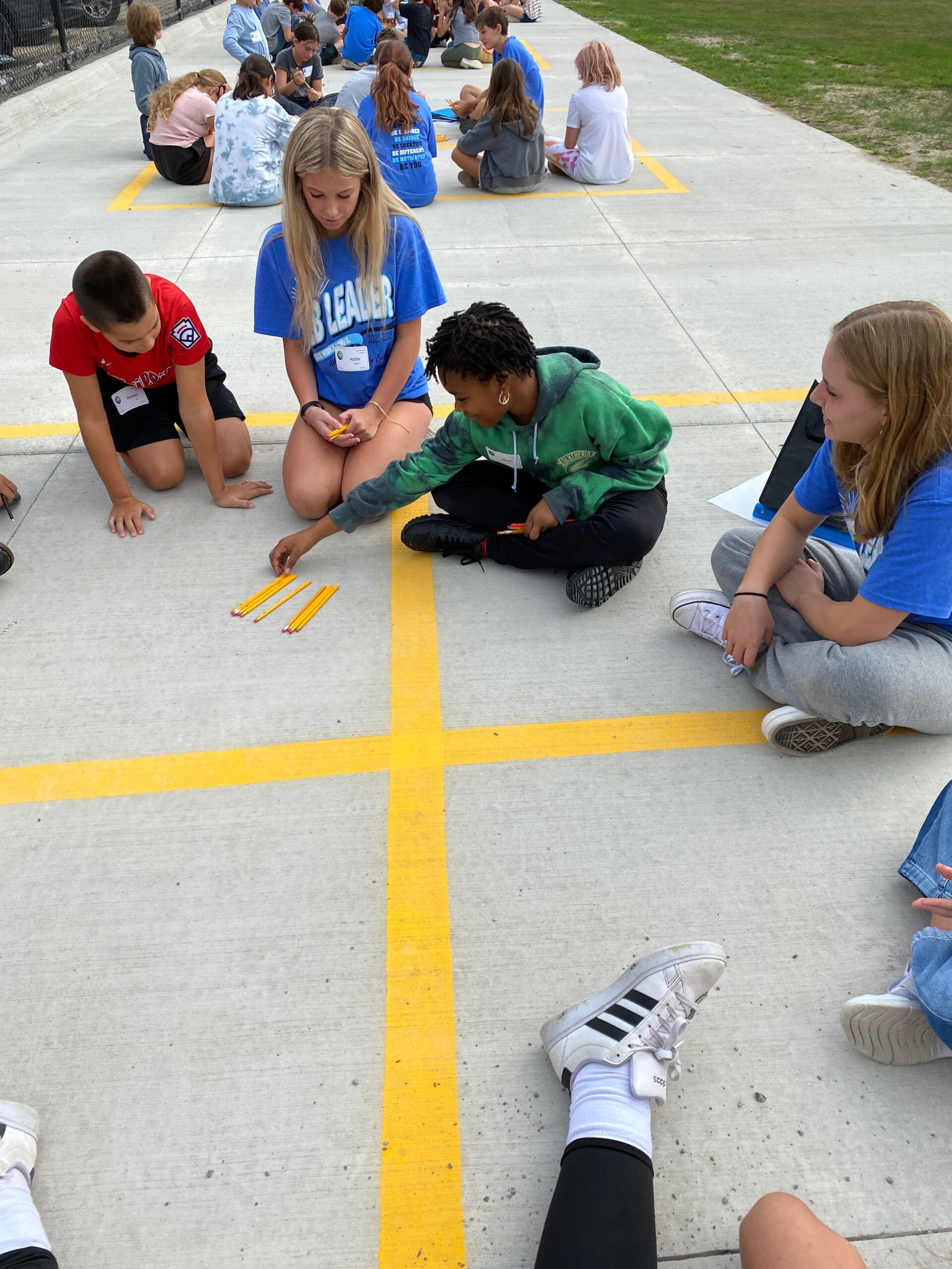 A small group of students sit on a concrete surface with yellow grid lines, arranging pencils as part of an outdoor activity during a larger gathering.