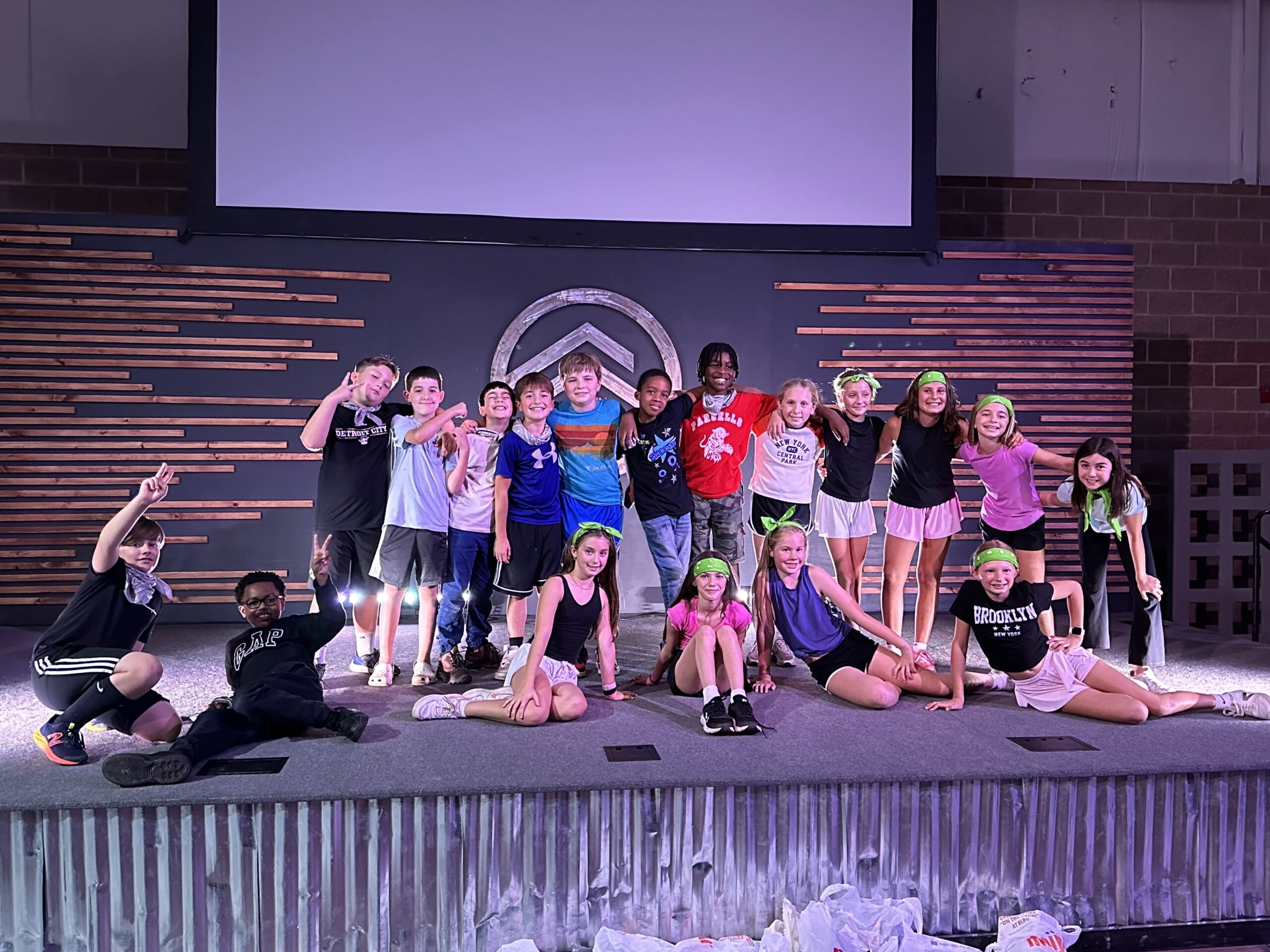 A group of children pose and smile together on an indoor stage, some wearing green bandanas, in front of a wooden-slat backdrop and large blank screen.