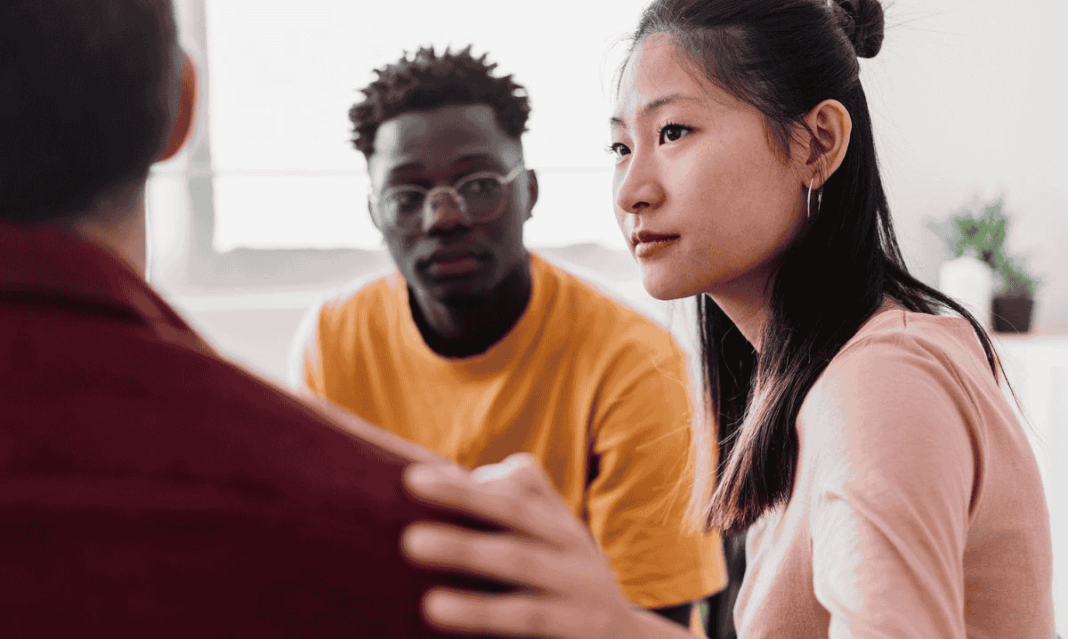 Three young adults sitting in a support group setting. One woman rests a reassuring hand on another person’s shoulder while listening attentively.