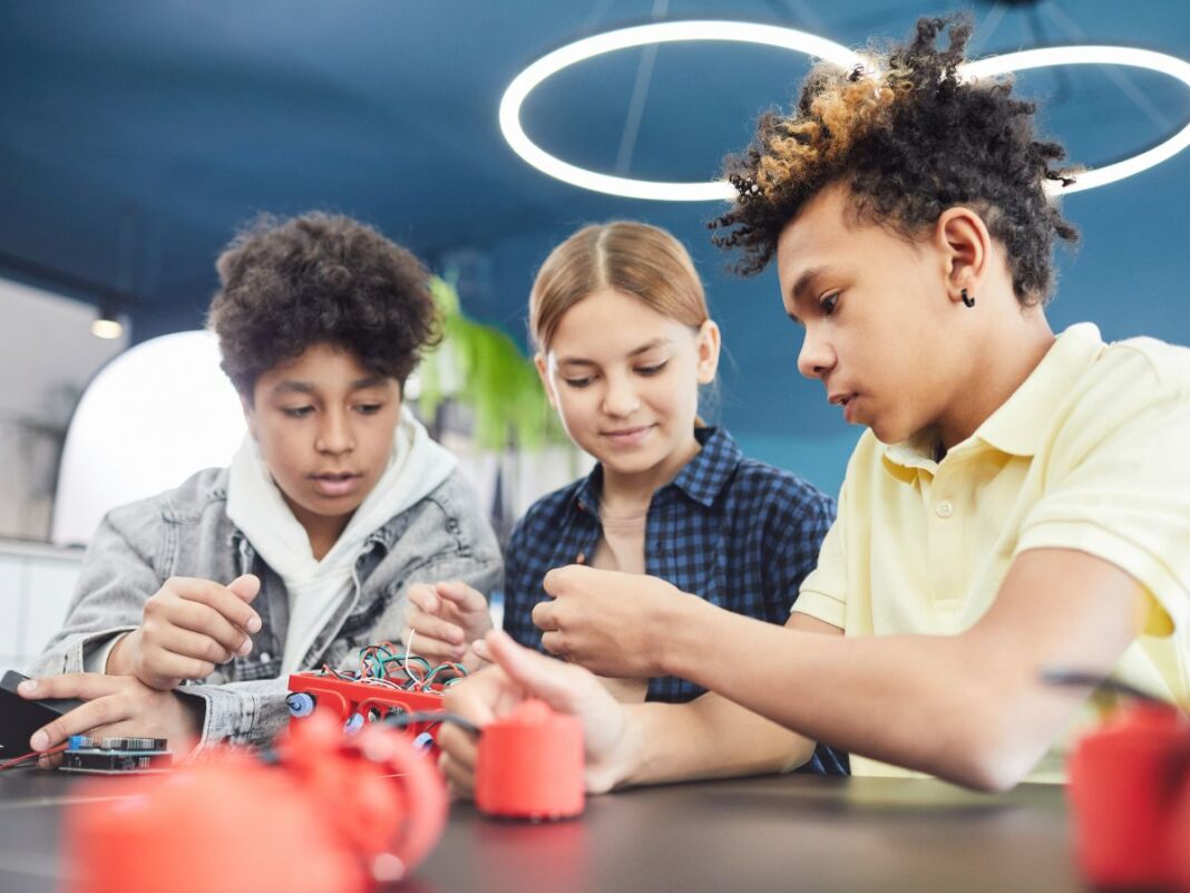 Three middle school students work together on a hands-on robotics project, focused on assembling red plastic components and wires. They are seated at a table in a brightly lit classroom with a modern, circular light fixture above them.