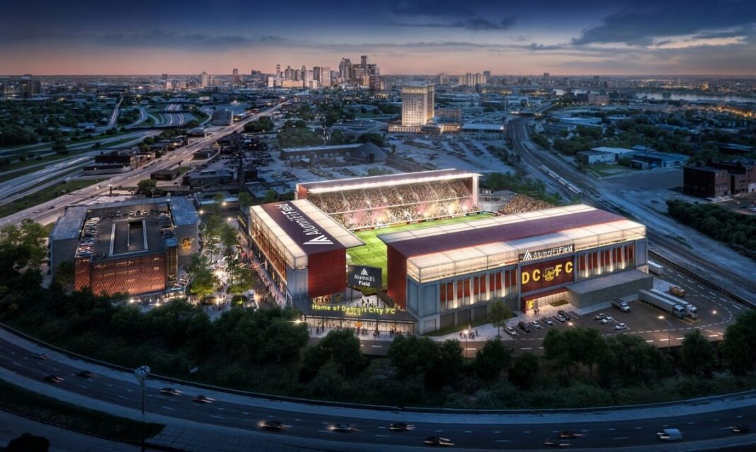 Aerial view of Detroit City FC’s new AlummiFi Field stadium at night, with lights, fans, and downtown Detroit skyline in the background.