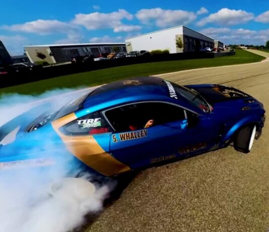 A blue drift car races at M1 Concourse during the American Speed Festival with smoke trailing from the tires on the track.