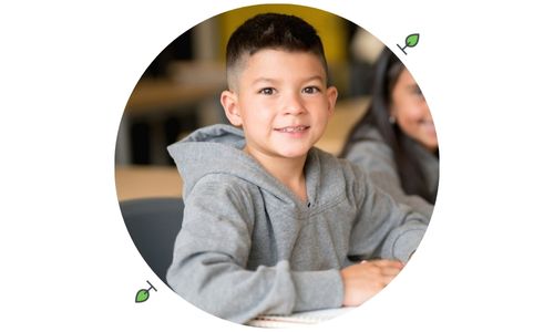 Smiling elementary school boy in a gray hoodie sitting at a classroom desk with a notebook, symbolizing back-to-school parent teacher communication for kids with autism.