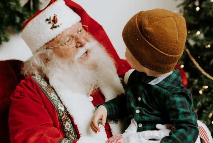 A young child wearing a brown hat and green shirt sits with Santa Claus, sharing a joyful holiday moment by the Christmas tree.