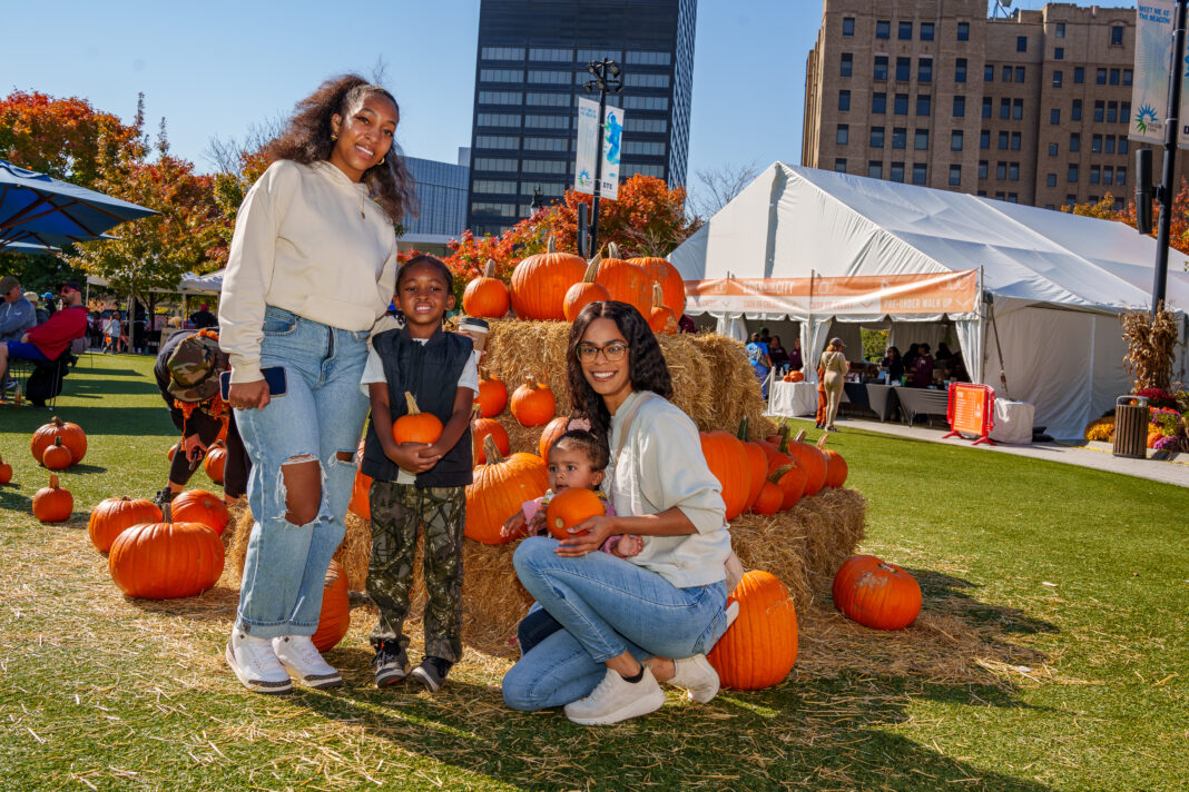 A smiling family poses in front of a pumpkin and haystack display at an outdoor fall festival. The mother stands beside her son holding a pumpkin, while another woman kneels with a toddler holding a small pumpkin.