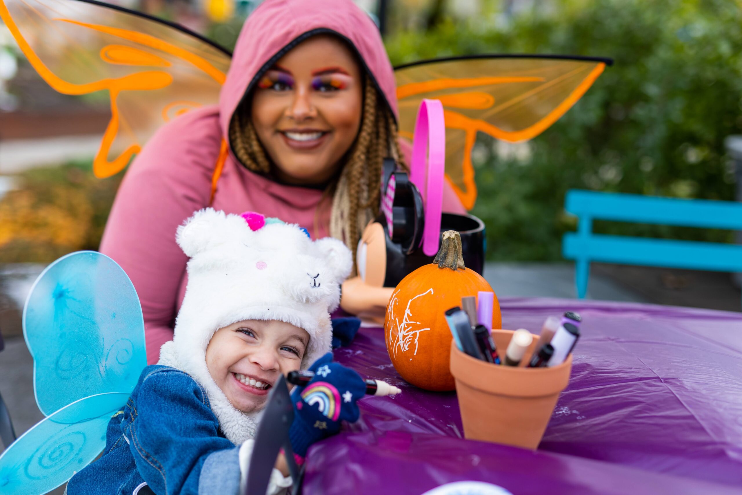 A child wearing fairy wings and a fuzzy animal hat smiles at a table while decorating a pumpkin. Behind them, an adult in butterfly wings looks on with a big smile.