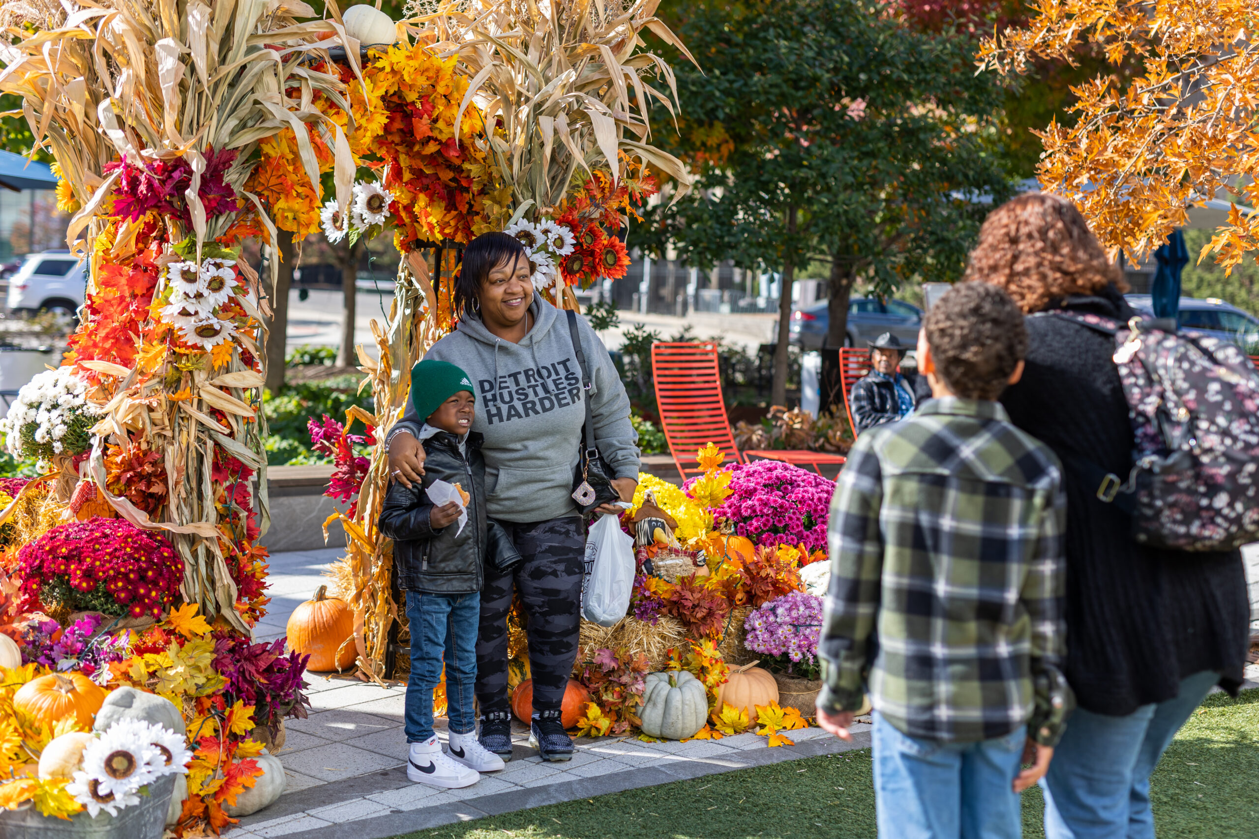 A woman in a hoodie with “Detroit Hustles Harder” printed on it poses with her son in front of a colorful autumn display featuring pumpkins, flowers, and cornstalks, while another family takes their photo.