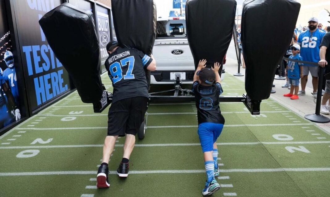 A father and son wearing Detroit Lions jerseys push tackling pads at the Ford truck push area in Pride Plaza before a football game.
