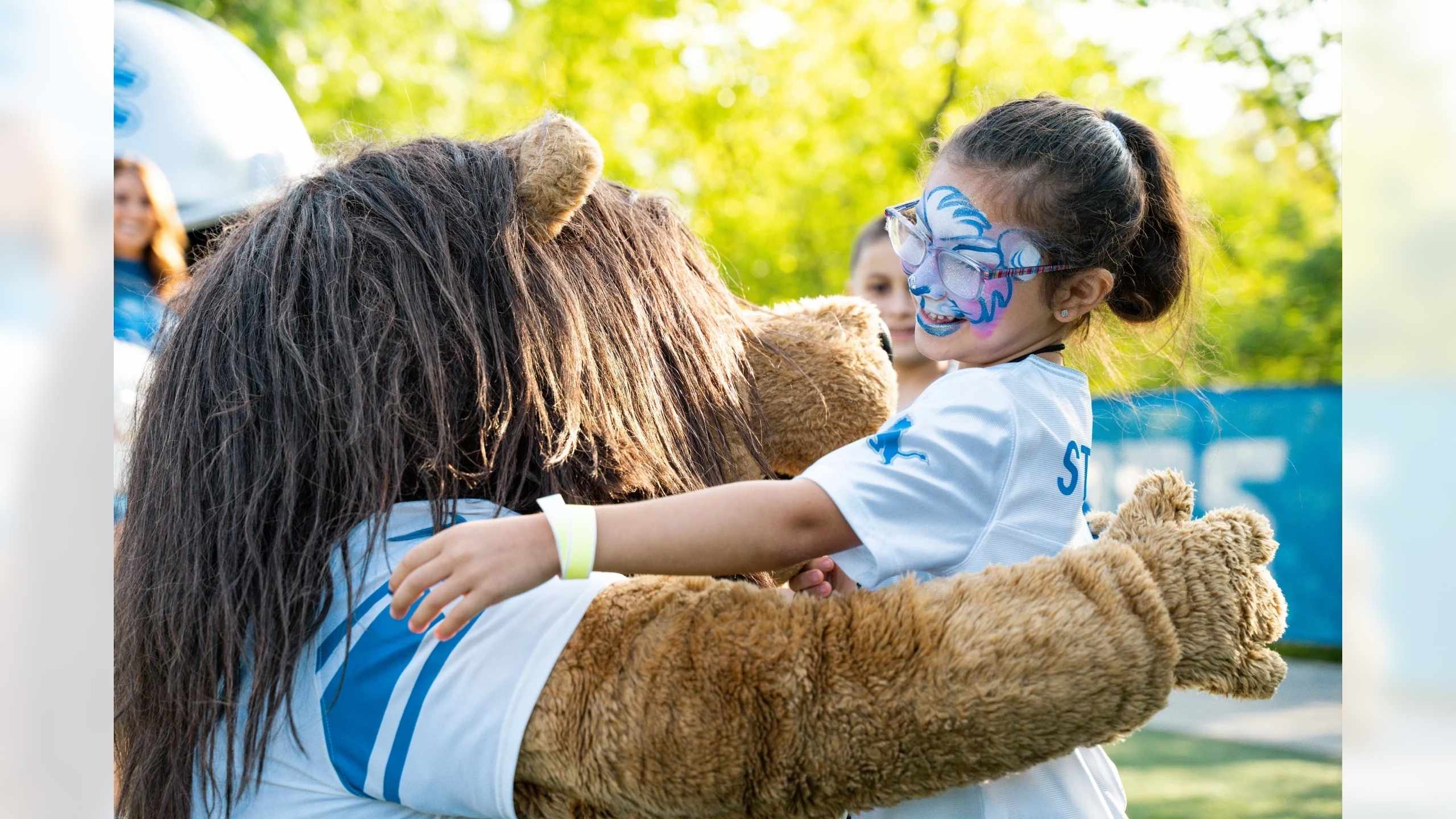 Young girl with face paint smiles while hugging Roary, the Detroit Lions mascot, during a Kids Club event filled with family fun
