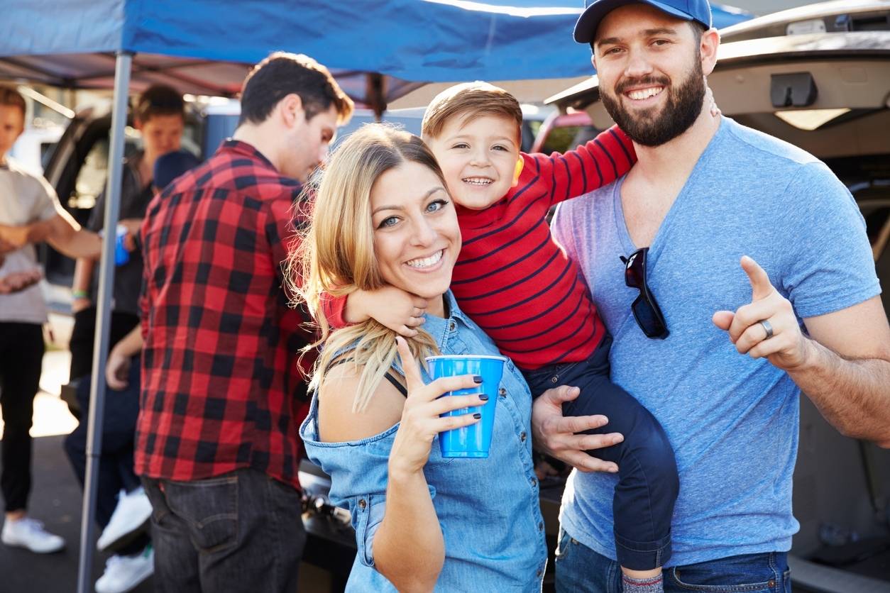 A smiling family with a young child enjoys tailgating before a football game in the parking lot with food and drinks.