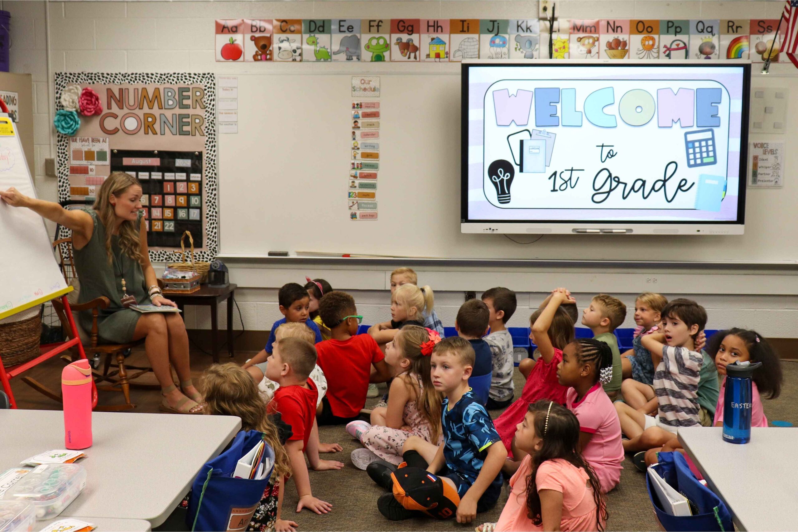 Farmington Public Schools first grade students sit on the floor while their teacher leads a lesson in a colorful classroom setting.