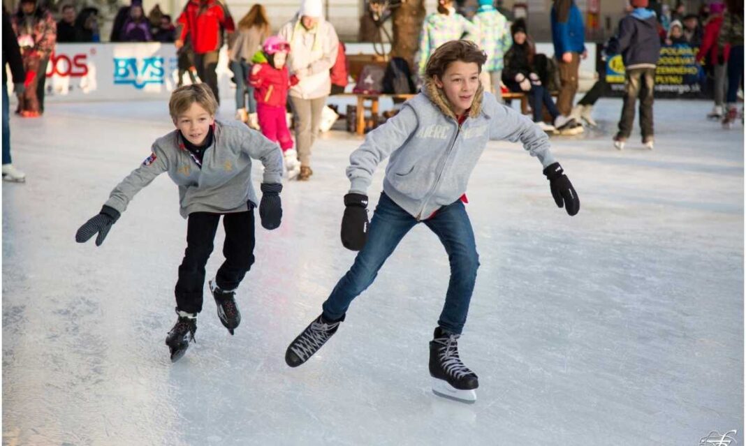 Two kids wearing gray jackets and gloves skate on an outdoor ice rink during winter while other families enjoy the activity in the background.