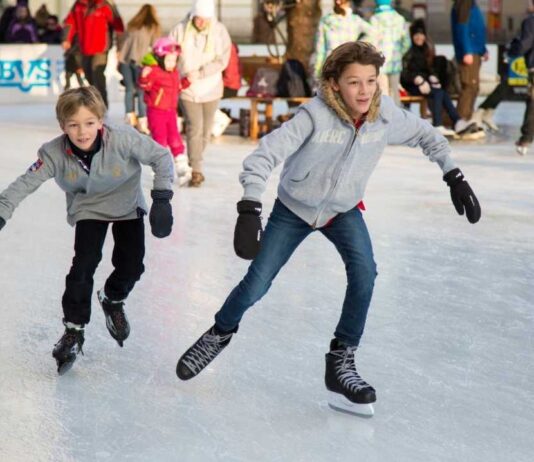 Two kids wearing gray jackets and gloves skate on an outdoor ice rink during winter while other families enjoy the activity in the background.