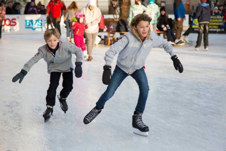 Two kids wearing gray jackets and gloves skate on an outdoor ice rink during winter while other families enjoy the activity in the background.