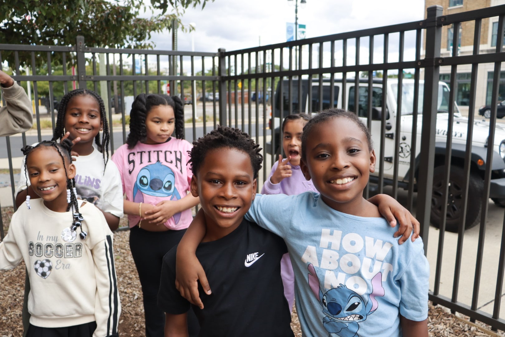 Group of smiling kids outside, two friends with arms around each other, wearing Stitch shirts and casual clothes by a fence