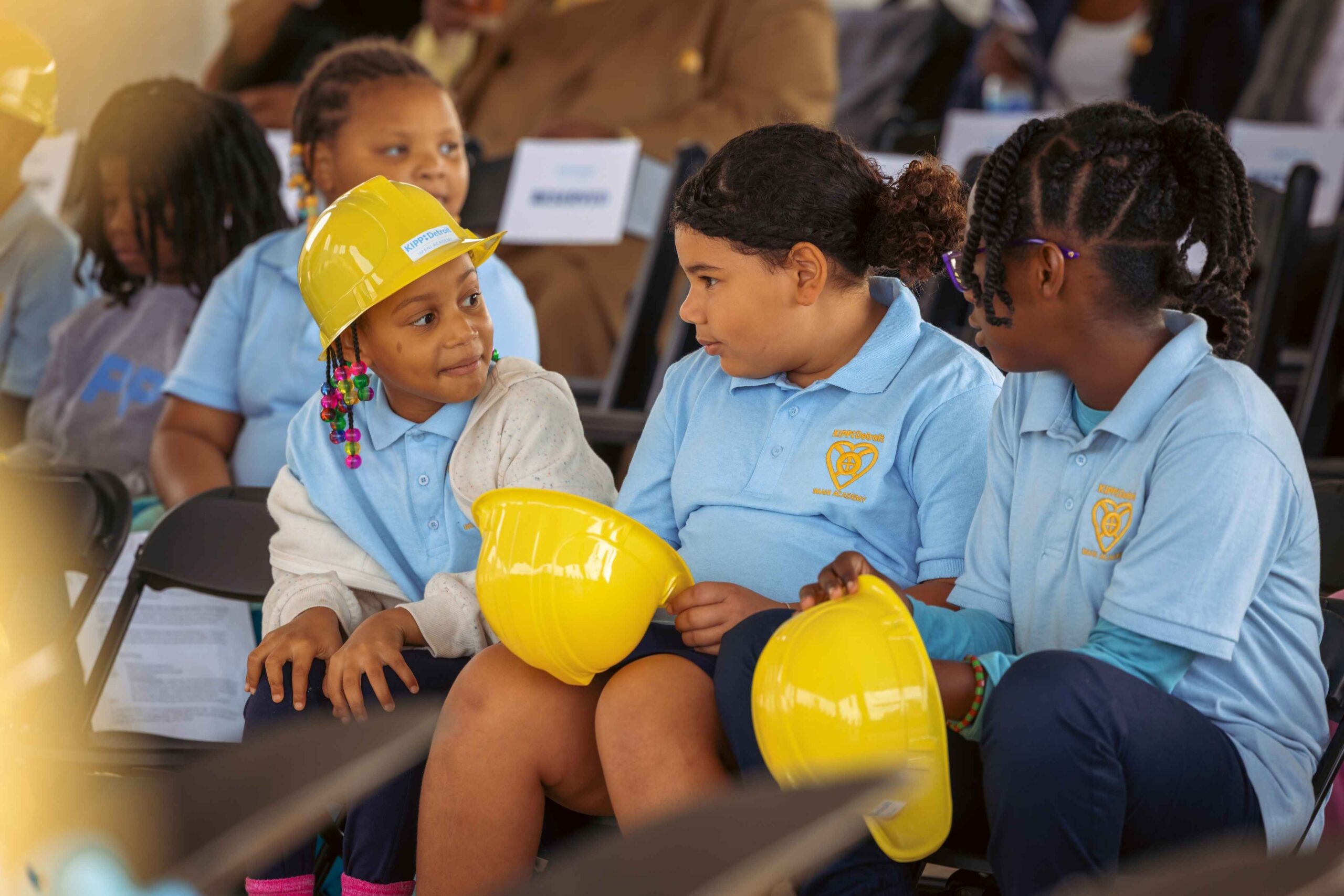 KIPP Detroit Charter School students in blue uniforms sit together holding yellow hard hats during a school event.