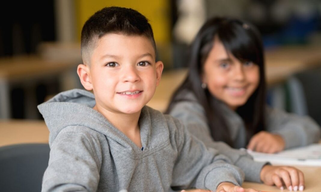 Two elementary school children smiling in a classroom, sitting at desks with notebooks and colored pencils — representing back-to-school and parent teacher communication support for kids with autism.