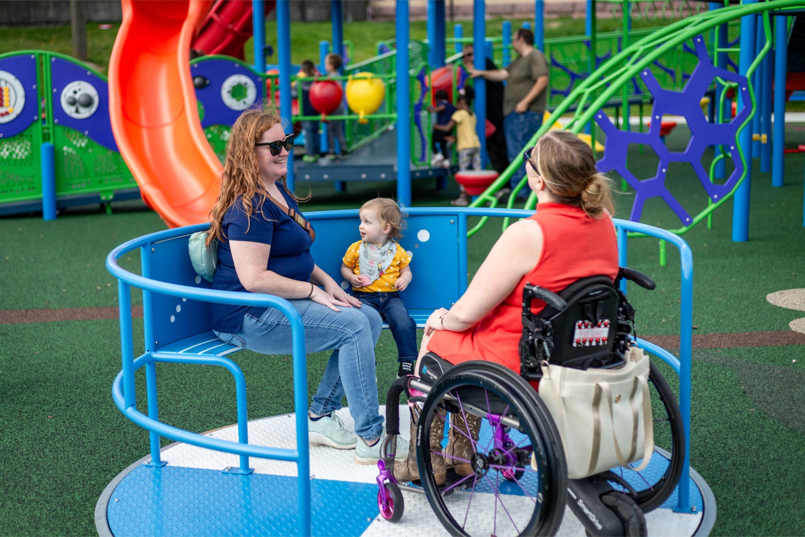 A mom in a wheelchair enjoys time with her child and friend on accessible play equipment at a Michigan playground with kids playing nearby.