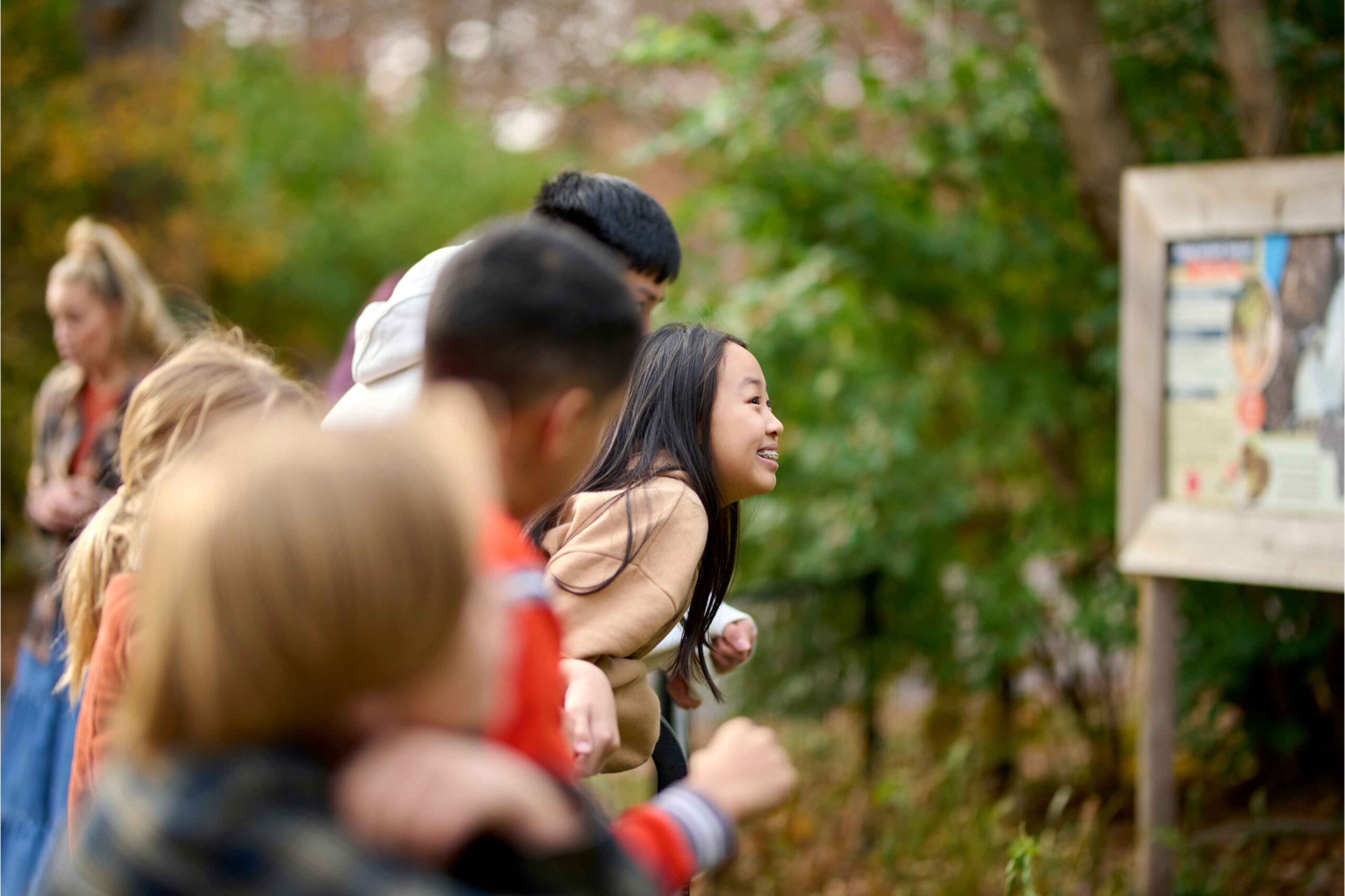 A group of kids lean over a railing at Potter Park Zoo reading an outdoor sign while learning about animals in a natural setting.