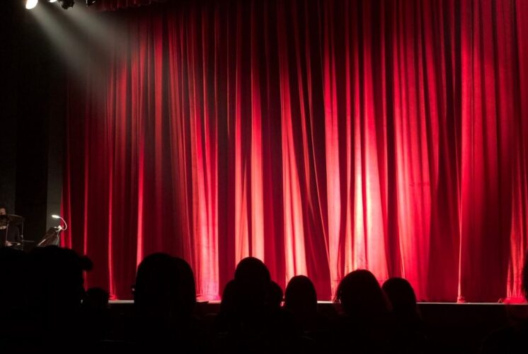Audience waits in front of red stage curtains before a live holiday musical performance of Rudolph the Red-Nosed Reindeer.
