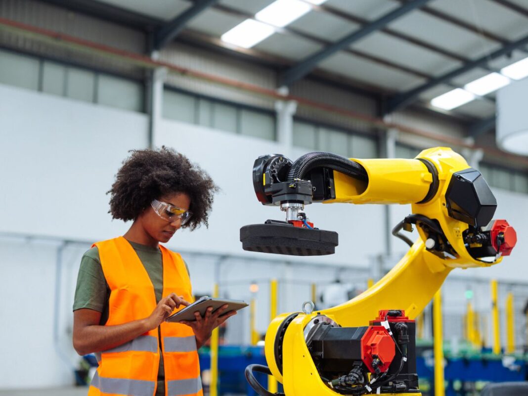 Student in safety vest using tablet to control industrial robotic arm during engineering training at Wayne County Community College District