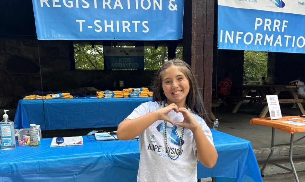 Aria LoVasco smiles and makes a heart with her hands at the Hope for Vision Walk, where her Michigan family raises awareness for rare eye diseases.