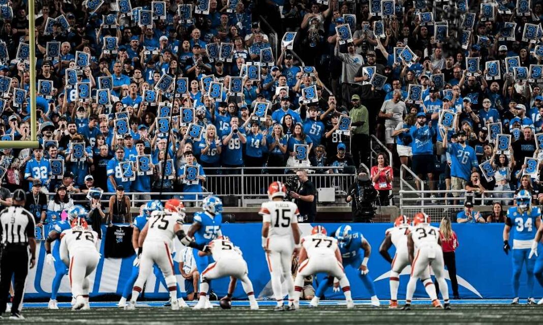 Detroit Lions fans fill the stands holding number three signs during a home game at Ford Field, showing team pride and game day excitement.
