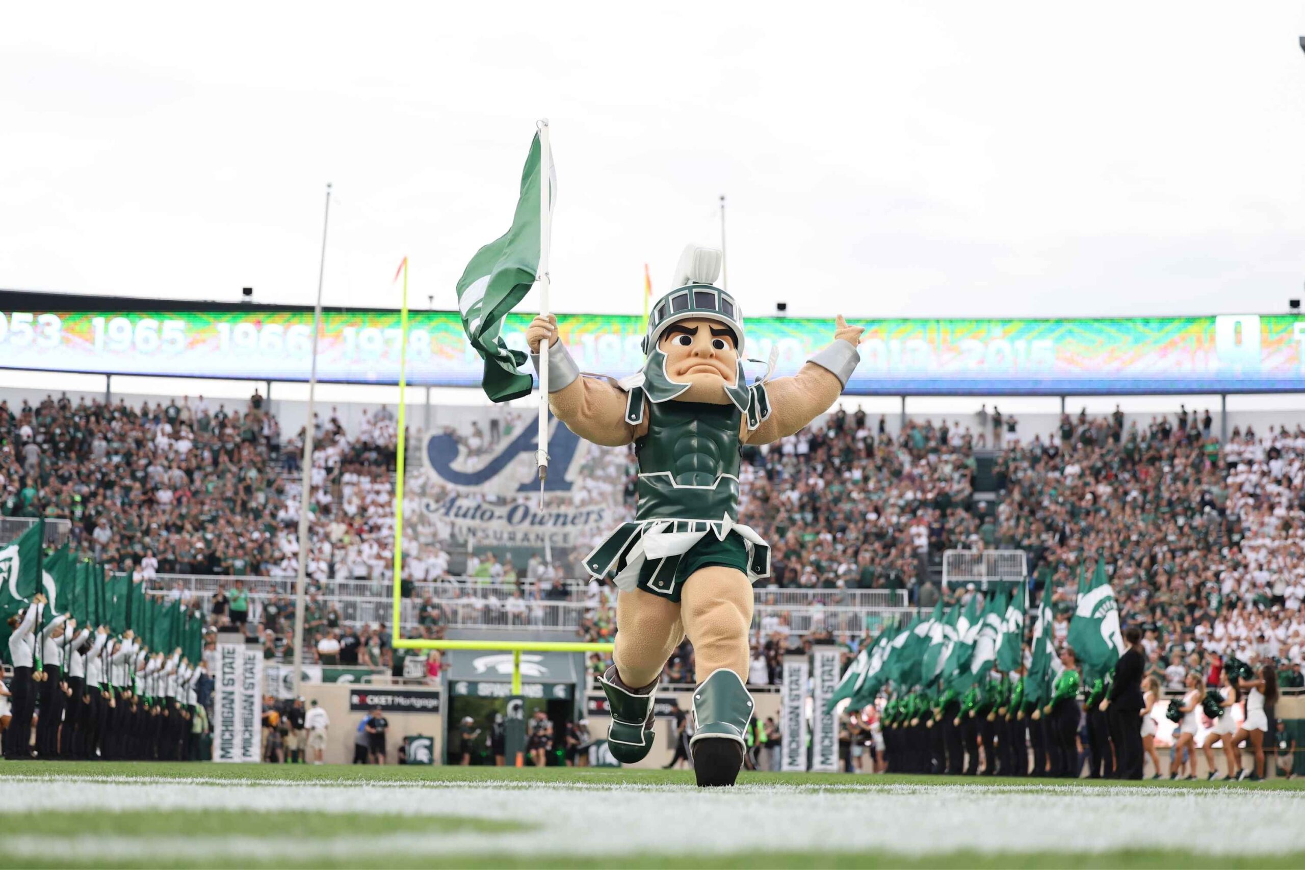 Michigan State University’s Sparty mascot runs onto the football field waving a green flag as fans cheer during a lively Spartans game day.