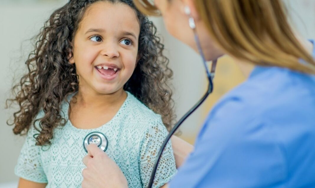 A young girl smiles as a doctor listens to her chest with a stethoscope, representing autism-informed care for children at Henry Ford Health.