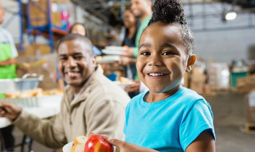 Child smiling while holding a plate of food and an apple at a community food pantry in Clinton Township.