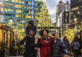 A family walks through the Downtown Detroit Markets with holiday lights and a large Christmas tree glowing behind them at Cadillac Lodge