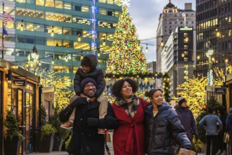 A family walks through the Downtown Detroit Markets with holiday lights and a large Christmas tree glowing behind them at Cadillac Lodge