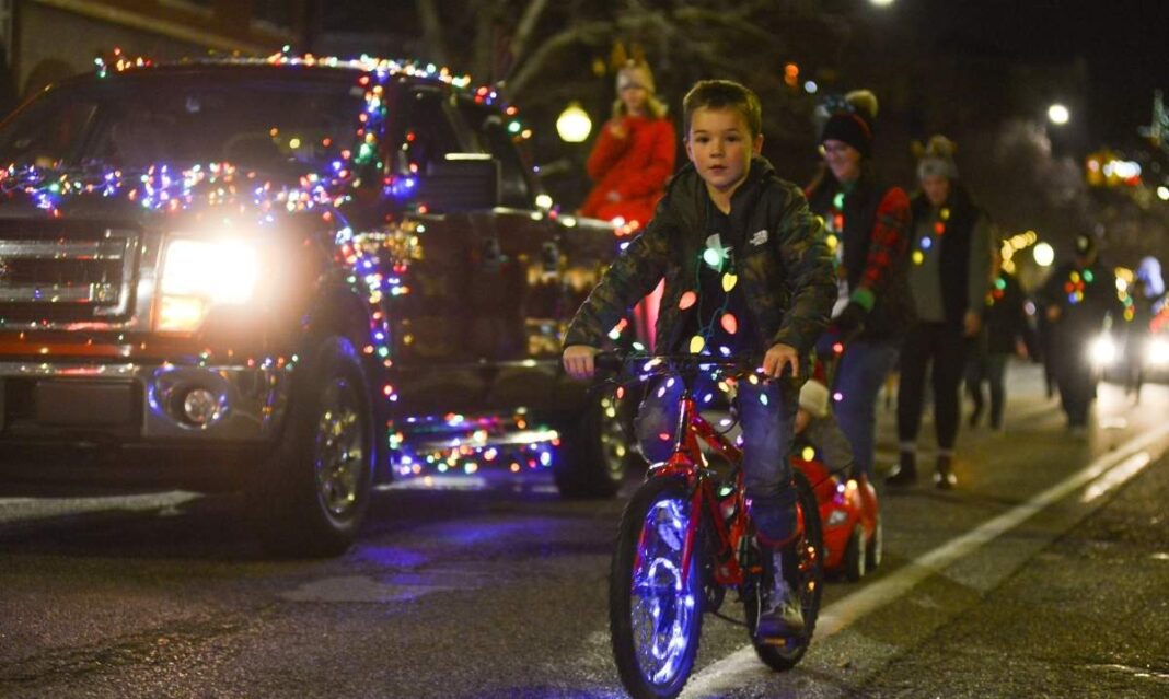 Child riding a bike decorated with holiday lights during the Holland Michigan Christmas parade with families walking behind and festive displays.