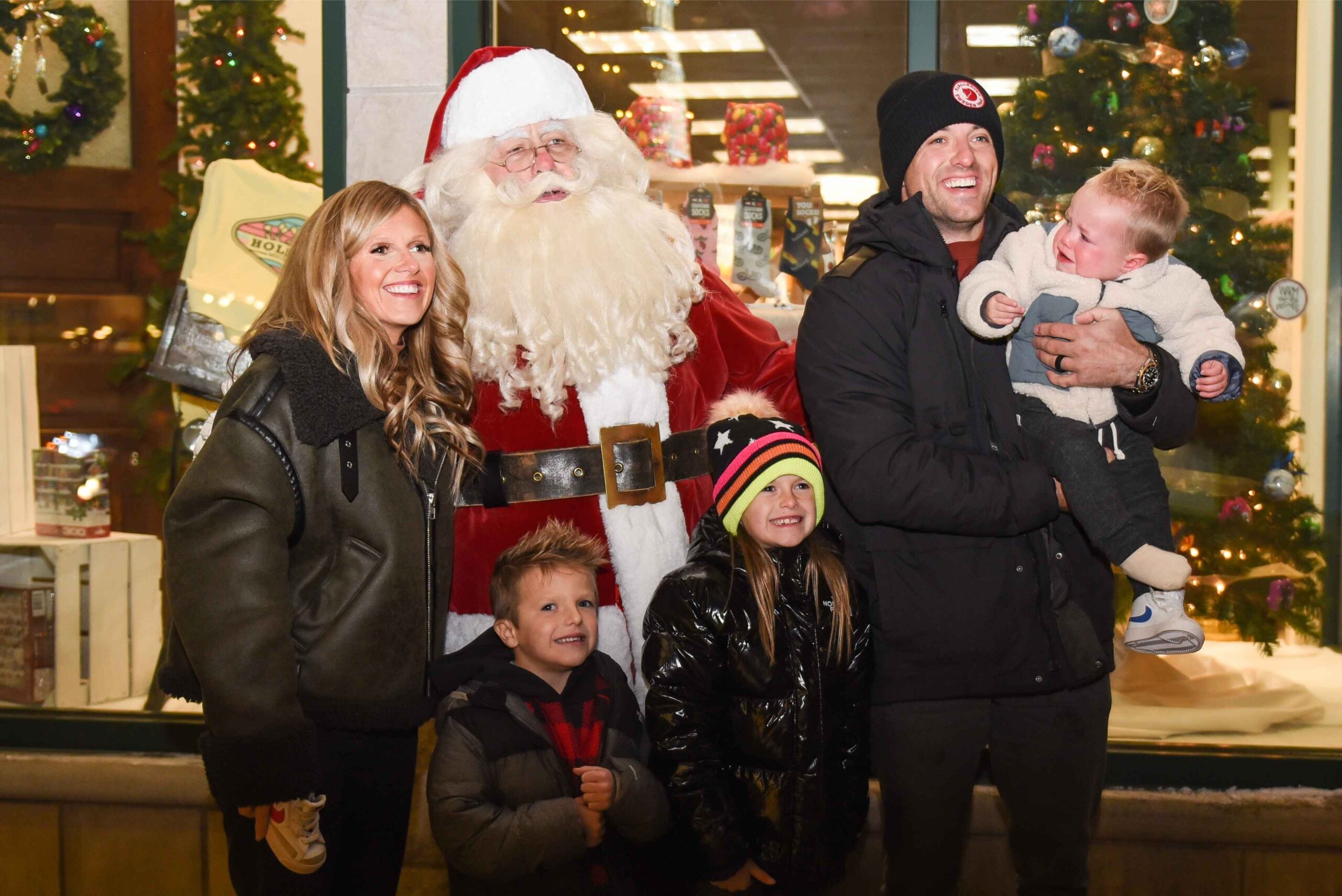 A family poses with Santa during a festive Holland Michigan Christmas event with holiday lights and decorations in the background.