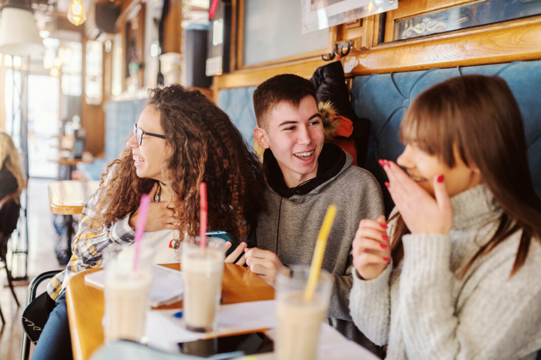 Three multicultural teenage classmates sitting in cafeteria and gossiping. On desk coffee, notebooks and pens.