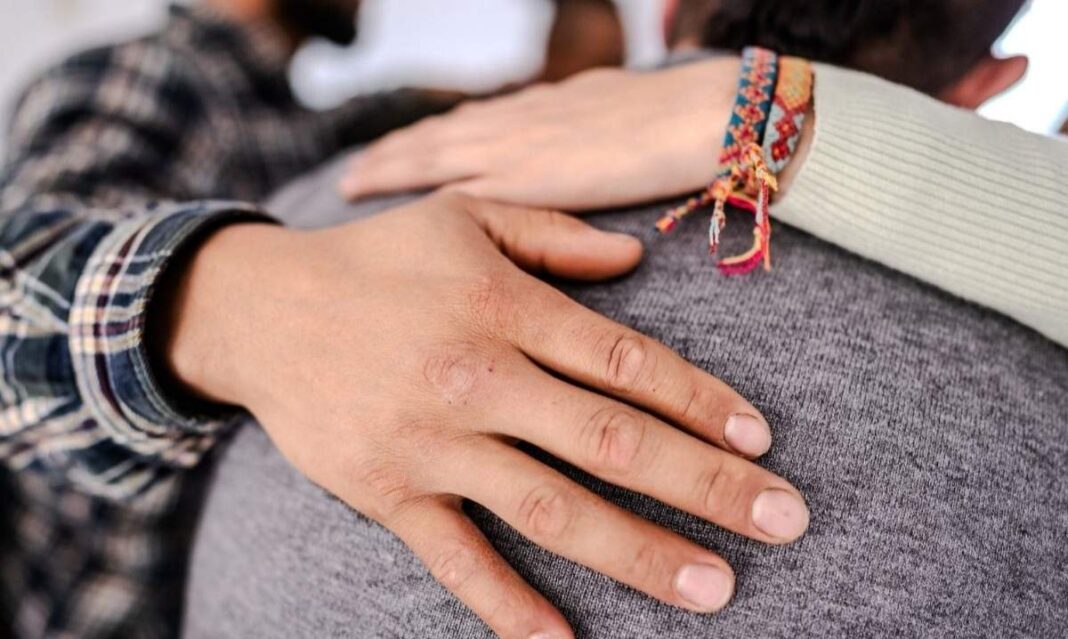 Close-up of supportive hands on a person’s shoulder during a peer recovery group session in metro Detroit