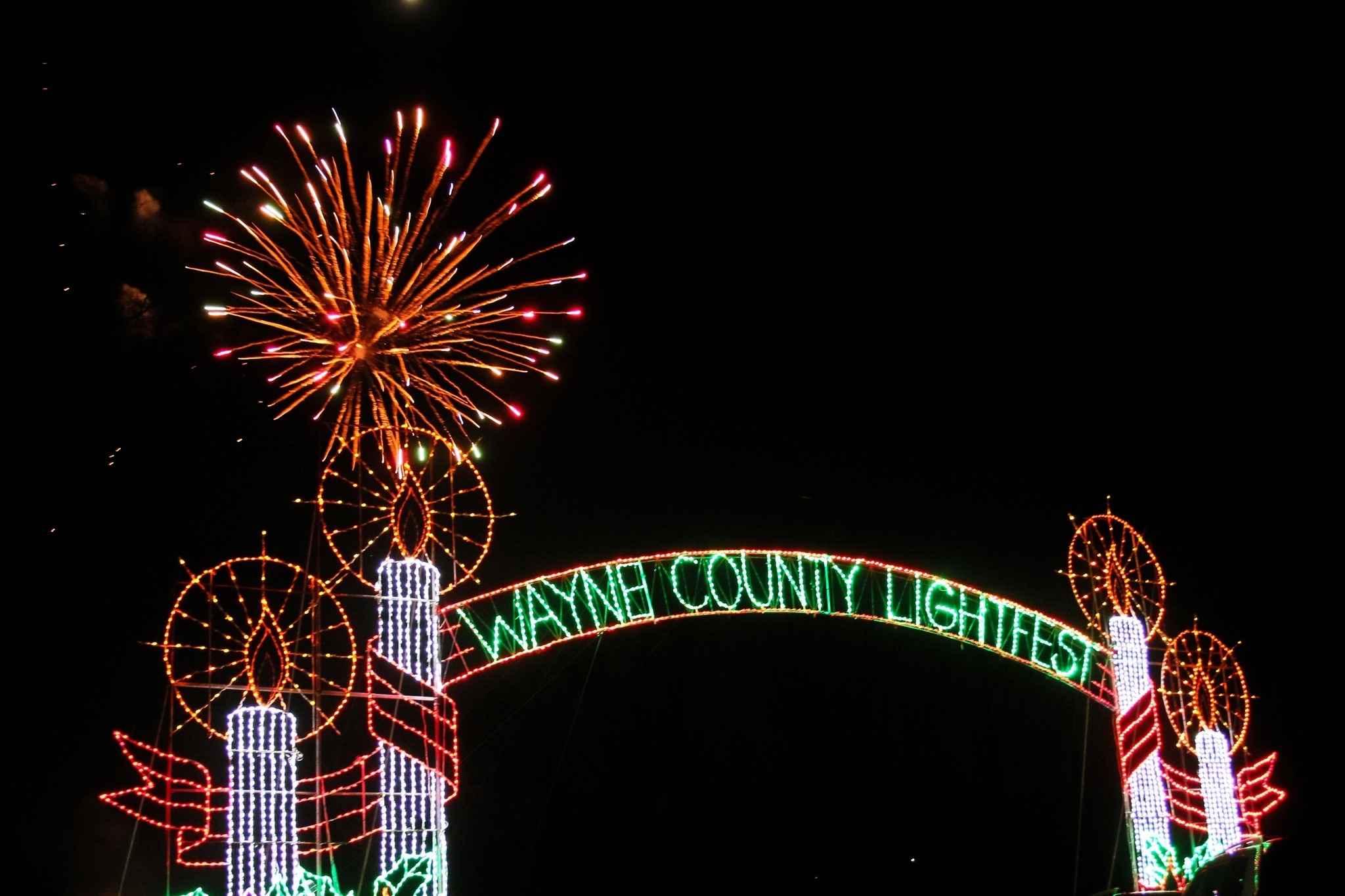 Fireworks burst above the Wayne County Lightfest entrance sign lit with candles and holiday lights in Westland Michigan.