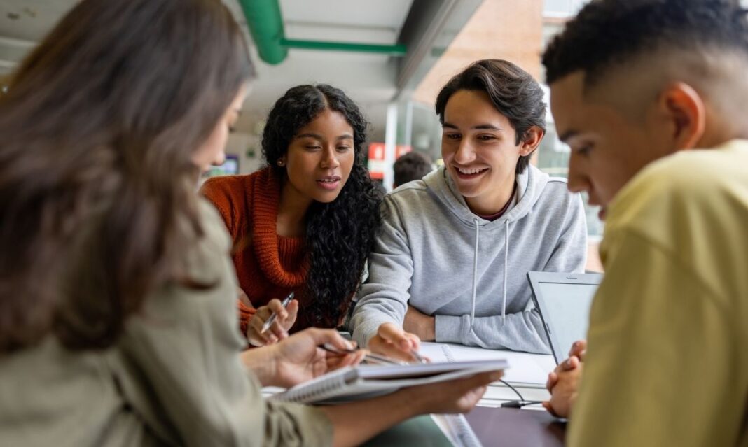A group of diverse high school students collaborate at a table, smiling and reviewing notes together, representing the supportive and engaging environment of the Early College of Macomb program.