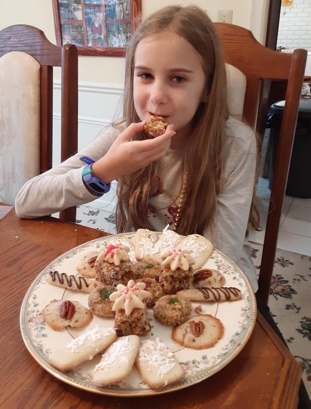 A girl enjoying a plate of her family’s traditional Christmas cookies made by her aunt Mary, sitting at the table with a full holiday cookie platter.