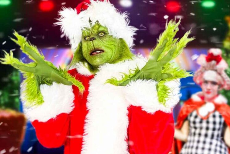 The Grinch in a Santa costume poses onstage during The Grinch And Cindy Lou Who Show at Fairytale Fun Place with festive lights in the background