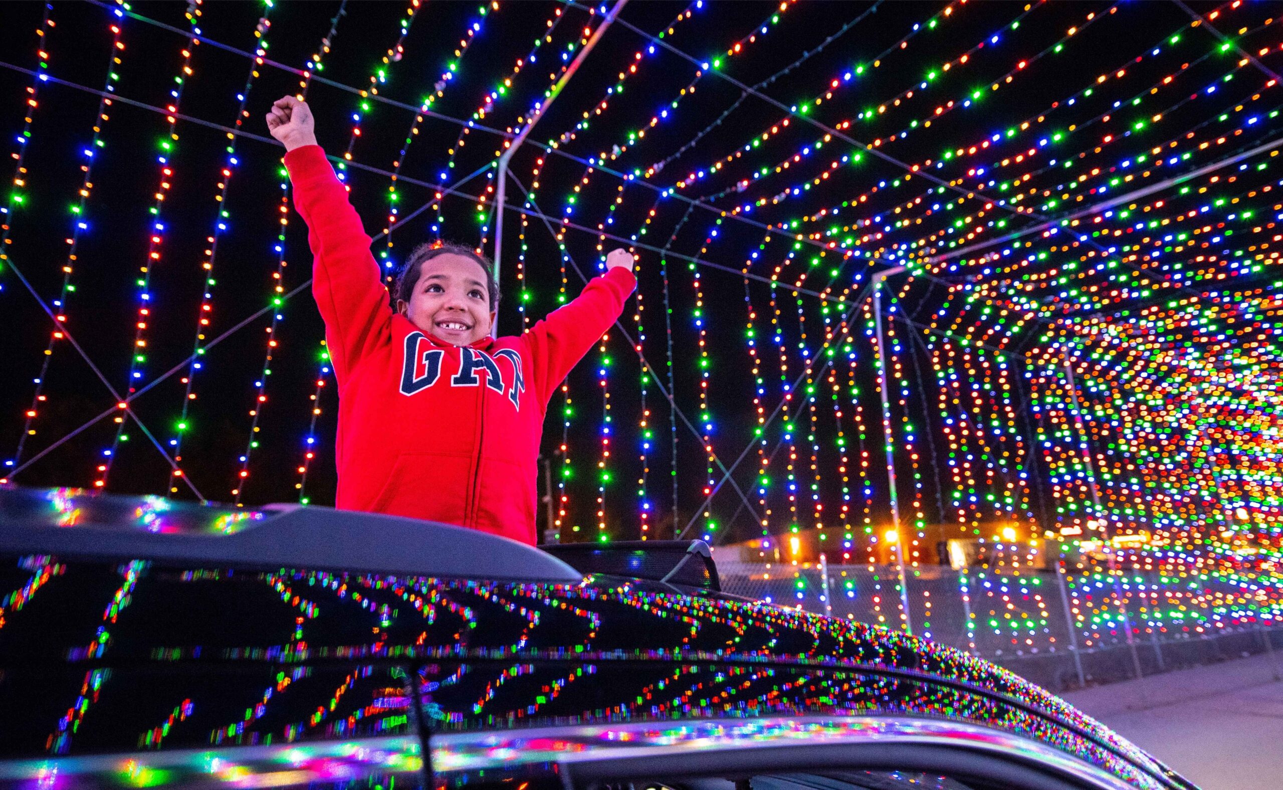 A child looks out of a car sunroof while driving through the Magic of Lights tunnel in Detroit showing colorful holiday lights above.