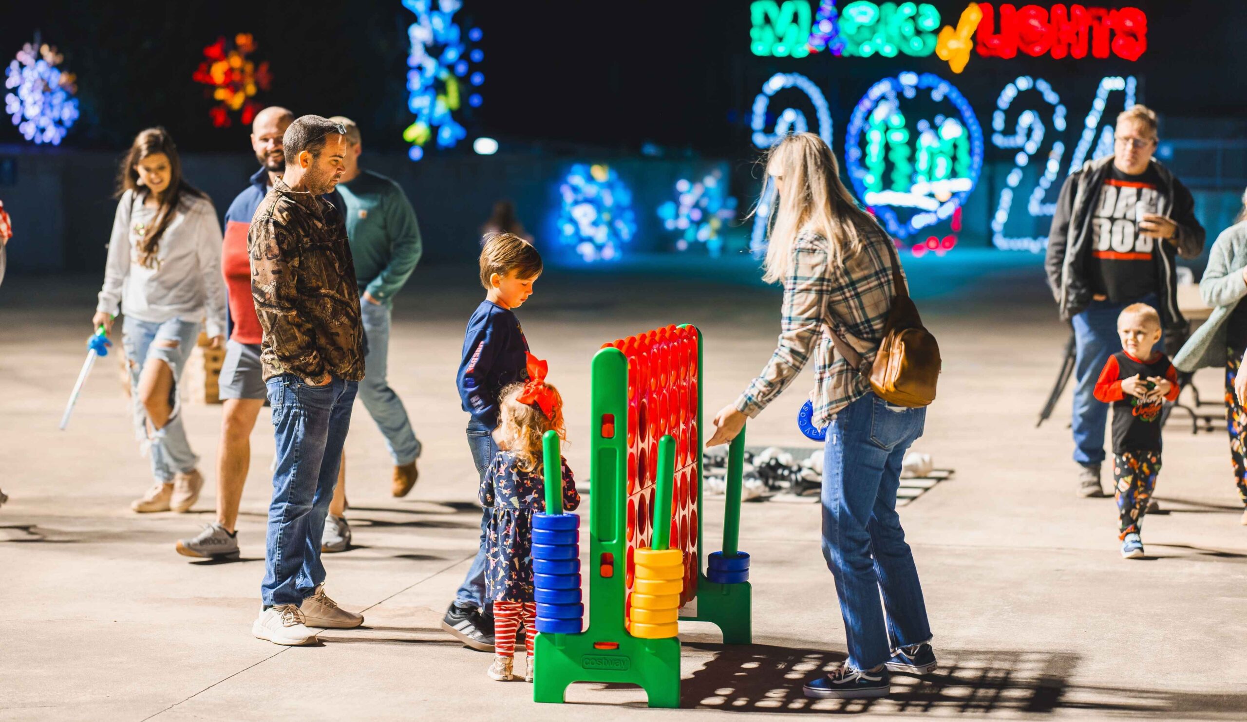 Families play games at the Magic of Lights walk-through area in Detroit showing kids enjoying activities beyond the drive-through display.