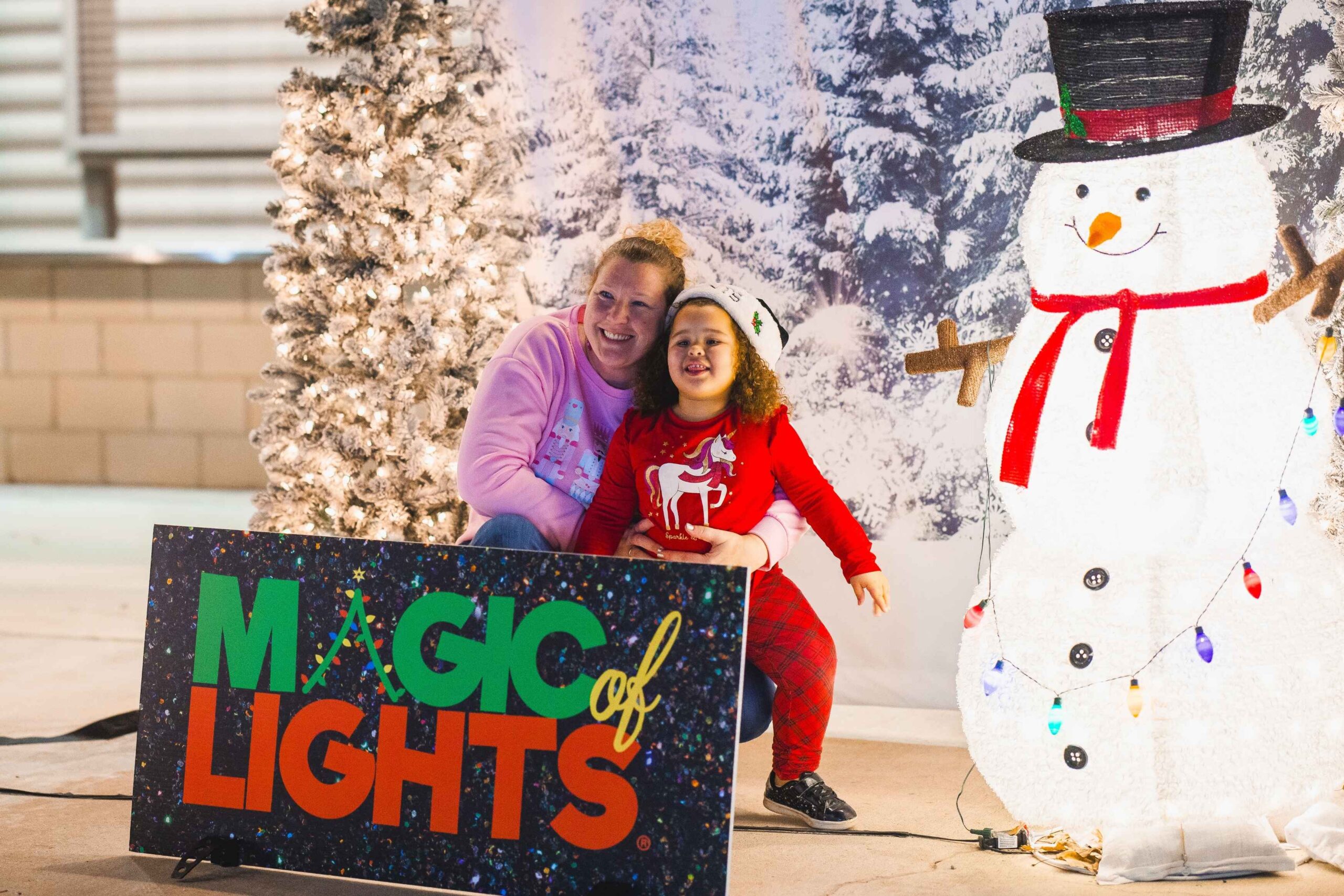 A parent and child pose beside a Magic of Lights sign and glowing snowman during the drive-through holiday show at Pine Knob in Clarkston Michigan.